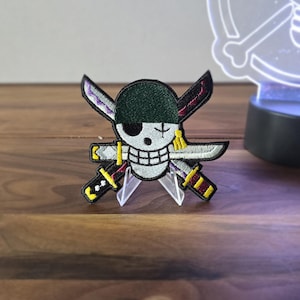 May include: Embroidered patch featuring a skull with a cross-shaped arrangement of three swords. The skull has a green eye patch and a scar. The swords have black, purple, and yellow details. The patch is displayed on a wooden surface.