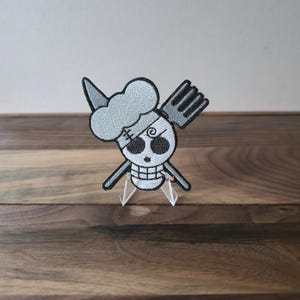 May include: Embroidered patch featuring a skull with a chef's hat and a fork, in shades of gray and black. The skull has an eye patch and a cross on its forehead. The patch is displayed on a wooden surface.