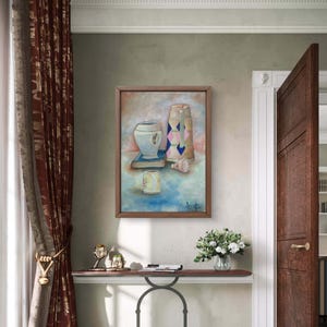 May include: A framed still life painting depicting a white vase, a tall patterned container, and a small cup. The artwork is in a wooden frame and hangs on a wall. The colour palette includes blues, pinks, and browns.