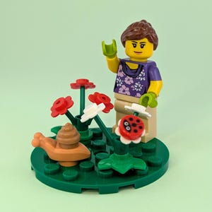 May include: A LEGO minifigure with brown hair wearing a purple floral top and tan pants, standing on a circular green base. The scene includes a brown snail, several red and white plastic flowers, and a small red ladybug piece on a stem.