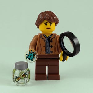Op de afbeelding: Een Lego-minifiguur met bruin haar, gekleed in een bruin jack en een bruine broek. De figuur houdt een vergrootglas in de ene hand en een klein, doorzichtig potje met een blauwe kever erin in de andere hand. De figuur houdt ook een klein, doorzichtig bakje met twee gele en zwarte bijen erin.