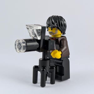 May include: A Lego minifigure depicting a photographer with black hair, a brown jacket, and yellow hands. The figure holds a black camera with a clear lens and flash, mounted on a black tripod. The background is plain white.