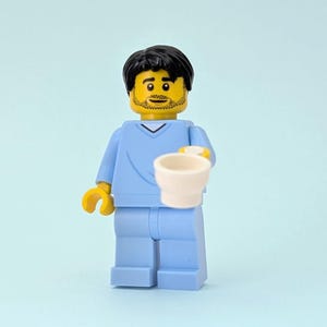 May include: A Lego figure in light blue scrubs, holding a white cup. The figure has black hair, a yellow face with a beard, and yellow hands. The background is a solid light blue.