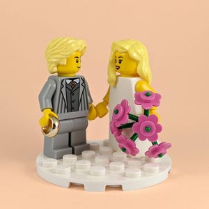 May include: A Lego wedding cake topper featuring a bride and groom. The groom wears a gray suit and holds a ring. The bride wears a white dress and holds a bouquet of pink flowers. The figures stand on a white base.