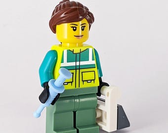 May include: A Lego minifigure with brown hair, a yellow head, and a green and yellow vest. The figure holds a light blue syringe and a white and gray object. The figure is wearing green pants and black gloves.