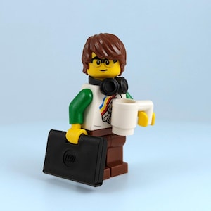 May include: A Lego minifigure with brown hair, glasses, and headphones. The figure wears a white shirt with a colourful design, green sleeves, and brown trousers. It holds a white mug and a black briefcase with the Lego logo.