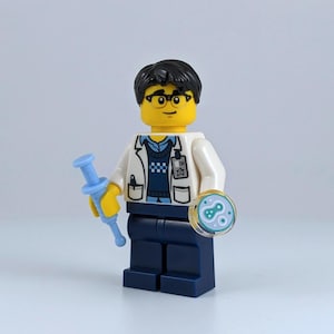 May include: A Lego minifigure of a scientist wearing a white lab coat, glasses, and dark hair. The figure holds a blue syringe in one hand and a petri dish with a blue and green design in the other. The scientist is wearing a blue sweater and dark blue pants.