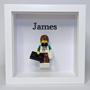 May include: A white square shadow box frame containing a Lego minifigure with brown hair, glasses, and a white shirt featuring an astronaut graphic. The figure holds a black briefcase and a white mug. The name James is printed in black above the figure.
