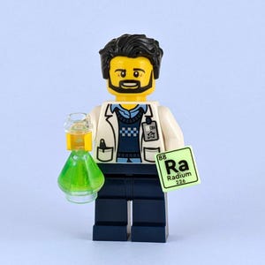 May include: A Lego scientist figure with a yellow head, black hair, and a beard, wearing a white lab coat and a blue sweater vest. The figure holds a green flask and a periodic table card with "Ra" for Radium.