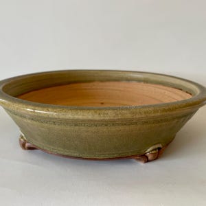 May include: A circular bonsai pot with a wide rim and three small feet. The pot is glazed in shades of green and brown, with a lighter interior. The design is simple and functional, suitable for bonsai cultivation.