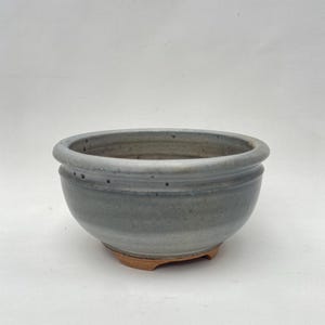 May include: A round, pale blue ceramic bonsai pot with a wide rim and a slightly textured surface. The pot has a small, unglazed base with three feet. The design is simple and functional.