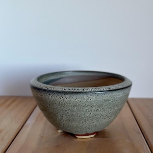 May include: A ceramic bowl with a speckled grey exterior and a dark rim. The bowl has a rounded shape and sits on three small feet. The interior is a warm brown colour. The bowl is on a wooden surface.