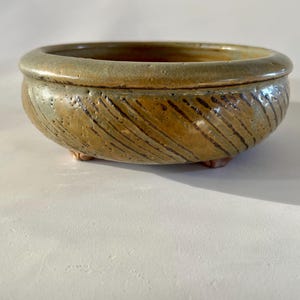 May include: A small, round ceramic bowl with a textured surface and a rim. The bowl is primarily tan with dark brown diagonal lines and rests on three small feet. The interior is a darker brown.