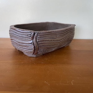 May include: A rectangular, brown ceramic bonsai pot with a textured, wave-like design. The pot has a dark brown interior and is set on a wooden surface. The pot's exterior features a unique, textured pattern.