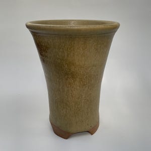 May include: An olive-green ceramic vase with a flared top and a textured surface. The vase has a unique, tapered shape and sits on a small, three-footed base. This decorative vase is suitable for displaying flowers or as a standalone art piece.