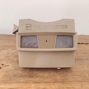 May include: A beige GAF View-Master stereoscope viewer featuring a rectangular body with two dark viewing lenses, a central ribbed panel, and a side-mounted advance lever. The brand name View-Master is embossed on the front top section.