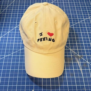 May include: A beige baseball cap with black embroidery that reads "I ♥ PEEING" with a red heart.