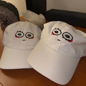 May include: Two white baseball caps with embroidered cartoon faces. The faces have black eyes, eyelashes, and eyebrows, red cheeks, and a black frown line.