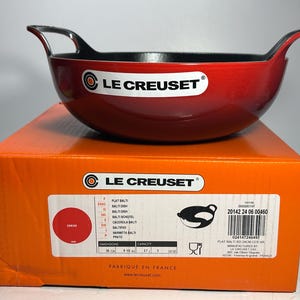 Könnte beinhalten: Eine rote Le Creuset Balti-Pfanne mit schwarzen Griffen, auf ihrer orangefarbenen Box. Die Pfanne hat einen Durchmesser von 24 cm. Die Box zeigt das Le Creuset-Logo und Produktinformationen.