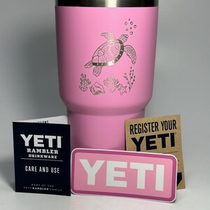 以下が含まれることがあります： ピンクのYETIランブラータンブラーに、ウミガメのデザインが施されています。タンブラーにはステンレス製の蓋と「YETI」と書かれたステッカーが付いています。