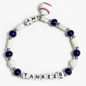 Op de afbeelding: Een kralenarmband met donkerblauwe en witte kralen, zilverkleurige tussenstukjes en witte letterkralen die YANKEES en NY spellen. Een kleine, ronde witte bedel met rode stiksels die op een honkbal lijkt, hangt aan de bovenkant van de armband.