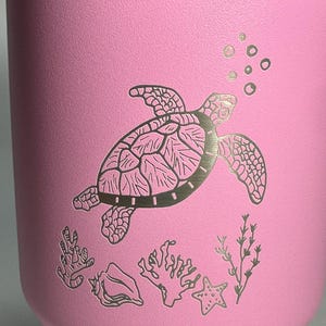 以下が含まれることがあります： ピンクのステンレス製のタンブラーに、レーザーエッチングで描かれた、泡、サンゴ、貝殻、ヒトデと一緒に泳ぐウミガメのデザインが施されています。