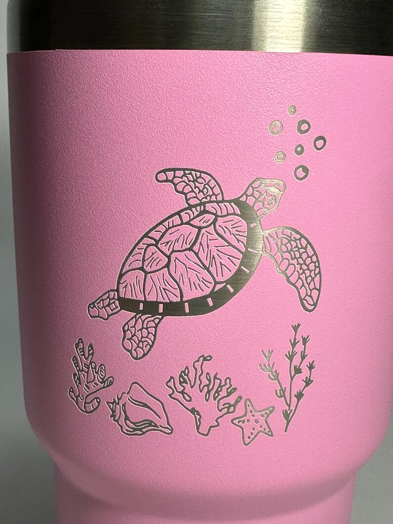以下が含まれることがあります： ピンクのステンレス製のタンブラーに、レーザーエッチングで描かれた、泡、サンゴ、貝殻、ヒトデと一緒に泳ぐウミガメのデザインが施されています。