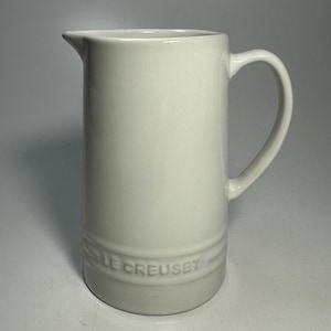 May include: A cream-coloured Le Creuset ceramic jug with a curved handle and a pouring spout. The jug has a cylindrical shape with a slightly flared base and the brand name embossed on the lower portion.