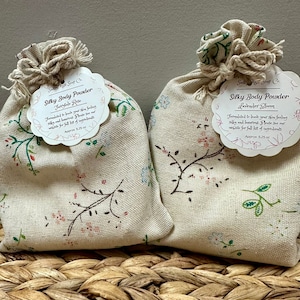 May include: Two small white fabric bags with floral patterns, tied with twine, containing silky body powder. The bags are labeled "Silky Body Powder" with the scents "Fairytale Rose" and "Lavender Bloom". The bags are sitting on a woven brown basket.