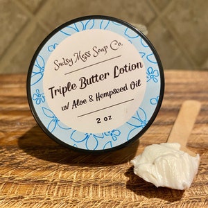 May include: A white jar with a blue lid and a label that reads "Sudsy Mess Soap Co. Triple Butter Lotion w/ Aloe & Hempseed Oil 2 oz". The jar is sitting on a wooden surface with a small dollop of lotion on a wooden spoon.