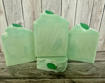 May include: Four square, light green soaps with a marbled design and a small green leaf on top. The soaps are arranged on a wooden surface with a wooden plank background. The soaps appear to be handmade.