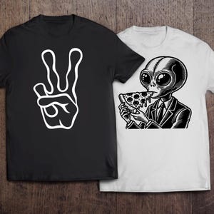 May include: Two black t-shirts with different designs. One shirt features a white peace sign with two fingers up. The other shirt features a black and white illustration of an alien eating a slice of pizza.