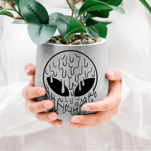 May include: A gray concrete planter with a black dripping alien face design. The planter is being held by a person's hands.