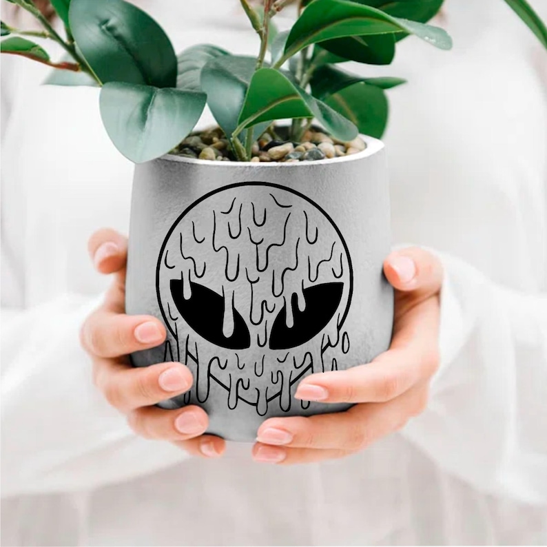 May include: A gray concrete planter with a black dripping alien face design. The planter is being held by a person's hands.