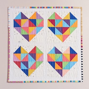 Pode incluir: Um edredom branco com quatro corações coloridos de patchwork. Cada coração é feito de uma variedade de cores brilhantes, incluindo vermelho, laranja, amarelo, verde, azul, roxo e rosa. O edredom é guarnecido com uma bainha colorida.