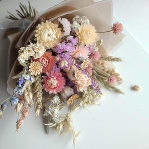 May include: A bouquet of dried flowers in shades of pink, peach, purple, and cream, wrapped in brown paper and tied with a ribbon. The arrangement includes various flower types and dried grasses, creating a rustic and natural aesthetic.