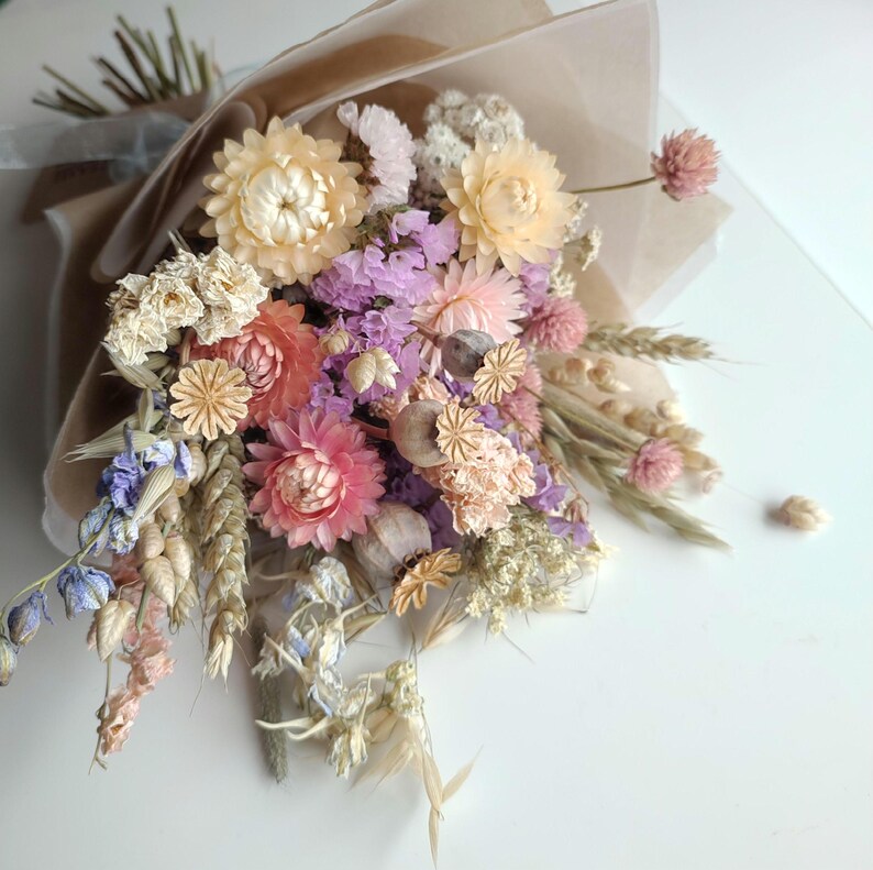 May include: A bouquet of dried flowers in shades of pink, peach, purple, and cream, wrapped in brown paper and tied with a ribbon. The arrangement includes various flower types and dried grasses, creating a rustic and natural aesthetic.