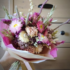 May include: A bouquet of dried flowers in shades of pink, purple, and cream, wrapped in pink and tan paper. The bouquet includes various flower types and grasses, tied with a pink ribbon and a tag.