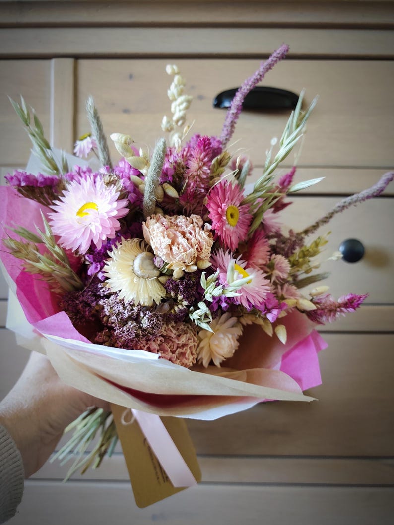 May include: A bouquet of dried flowers in shades of pink, purple, and cream, wrapped in pink and tan paper. The bouquet includes various flower types and grasses, tied with a pink ribbon and a tag.