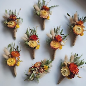 May include: A collection of seven dried flower buttonholes featuring yellow and orange strawflowers, fluffy beige bunny tails, and green foliage, each wrapped with natural twine at the base, arranged on a plain white background.