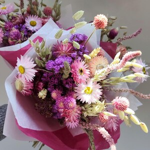 May include: A vibrant bouquet of dried flowers, featuring pink, purple, and yellow blooms. The arrangement includes daisies, globe amaranth, and other textured stems, wrapped in pink and white paper. A beautiful floral arrangement.
