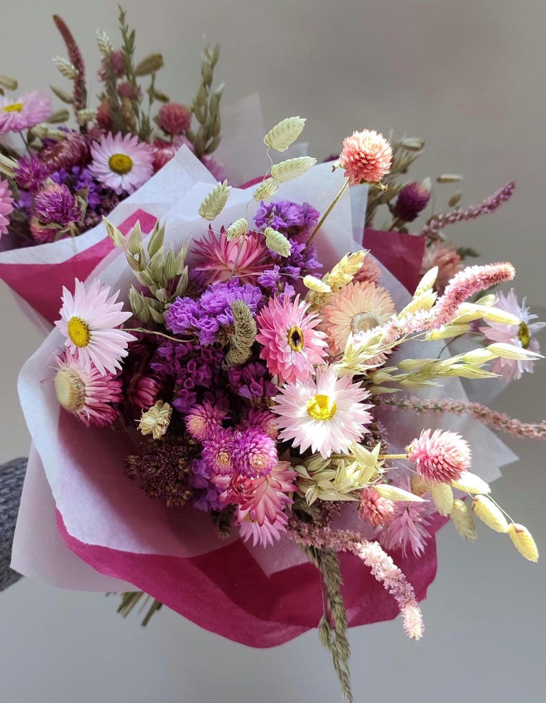 May include: A vibrant bouquet of dried flowers, featuring pink, purple, and yellow blooms. The arrangement includes daisies, globe amaranth, and other textured stems, wrapped in pink and white paper. A beautiful floral arrangement.