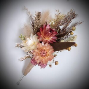 May include: A buttonhole featuring dried flowers including a deep red strawflower, a light peach strawflower, and small pink blossoms. The arrangement includes beige pampas grass, brown foliage, and textured seed pods, all wrapped in twine.