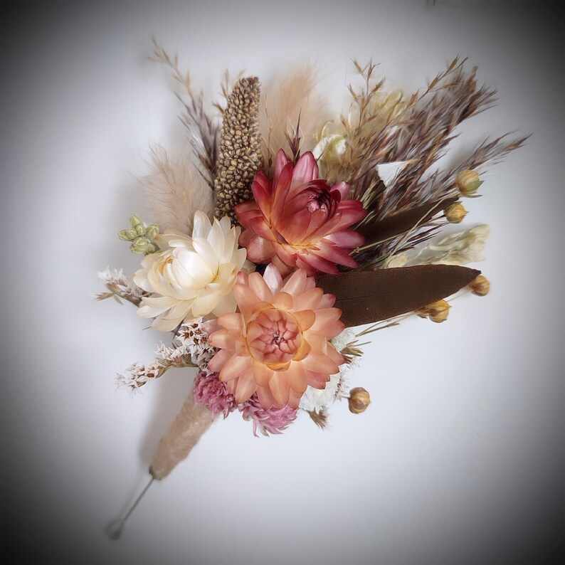 May include: A buttonhole featuring dried flowers including a deep red strawflower, a light peach strawflower, and small pink blossoms. The arrangement includes beige pampas grass, brown foliage, and textured seed pods, all wrapped in twine.