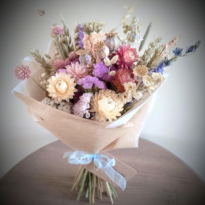 May include: A bouquet of dried flowers in shades of pink, purple, and cream, wrapped in brown paper. The bouquet is tied with a light blue ribbon and a small tag. The arrangement includes various flower types and wheat stalks.