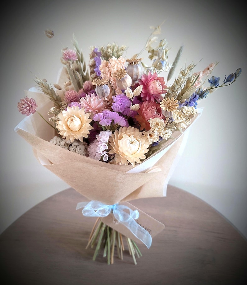 May include: A bouquet of dried flowers in shades of pink, purple, and cream, wrapped in brown paper. The bouquet is tied with a light blue ribbon and a small tag. The arrangement includes various flower types and wheat stalks.