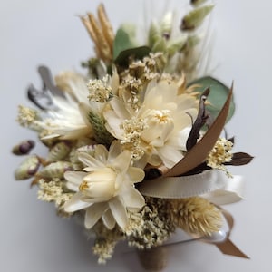 May include: A buttonhole featuring a collection of dried flowers in shades of cream, beige, and soft green. The arrangement includes small white blossoms, feathery grasses, and green leaves, accented with a thin cream ribbon and a brown ribbon tail.