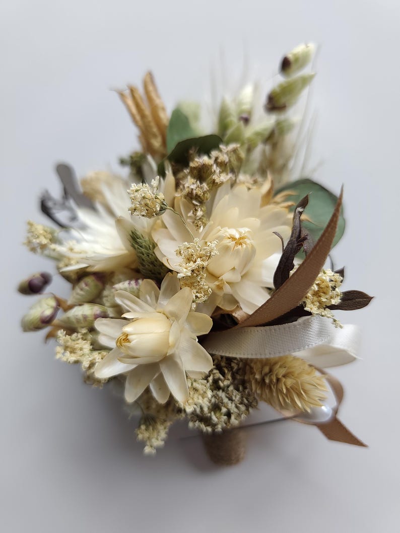 May include: A buttonhole featuring a collection of dried flowers in shades of cream, beige, and soft green. The arrangement includes small white blossoms, feathery grasses, and green leaves, accented with a thin cream ribbon and a brown ribbon tail.