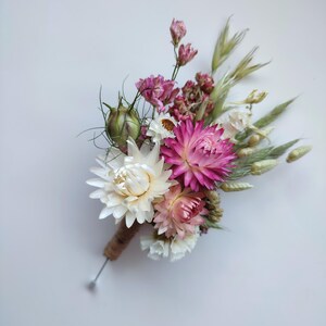 May include: A boutonniere featuring a collection of dried flowers including white and vibrant pink strawflowers, small pink buds, and green wheat stalks. The stems are bound together with brown twine, and a silver metal pin extends from the base.
