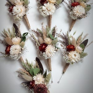 May include: A collection of seven buttonholes featuring dried flowers, including cream and burgundy strawflowers, fluffy pampas grass, green eucalyptus leaves, and wheat stalks. Each arrangement is wrapped in natural jute twine with a metal pin.